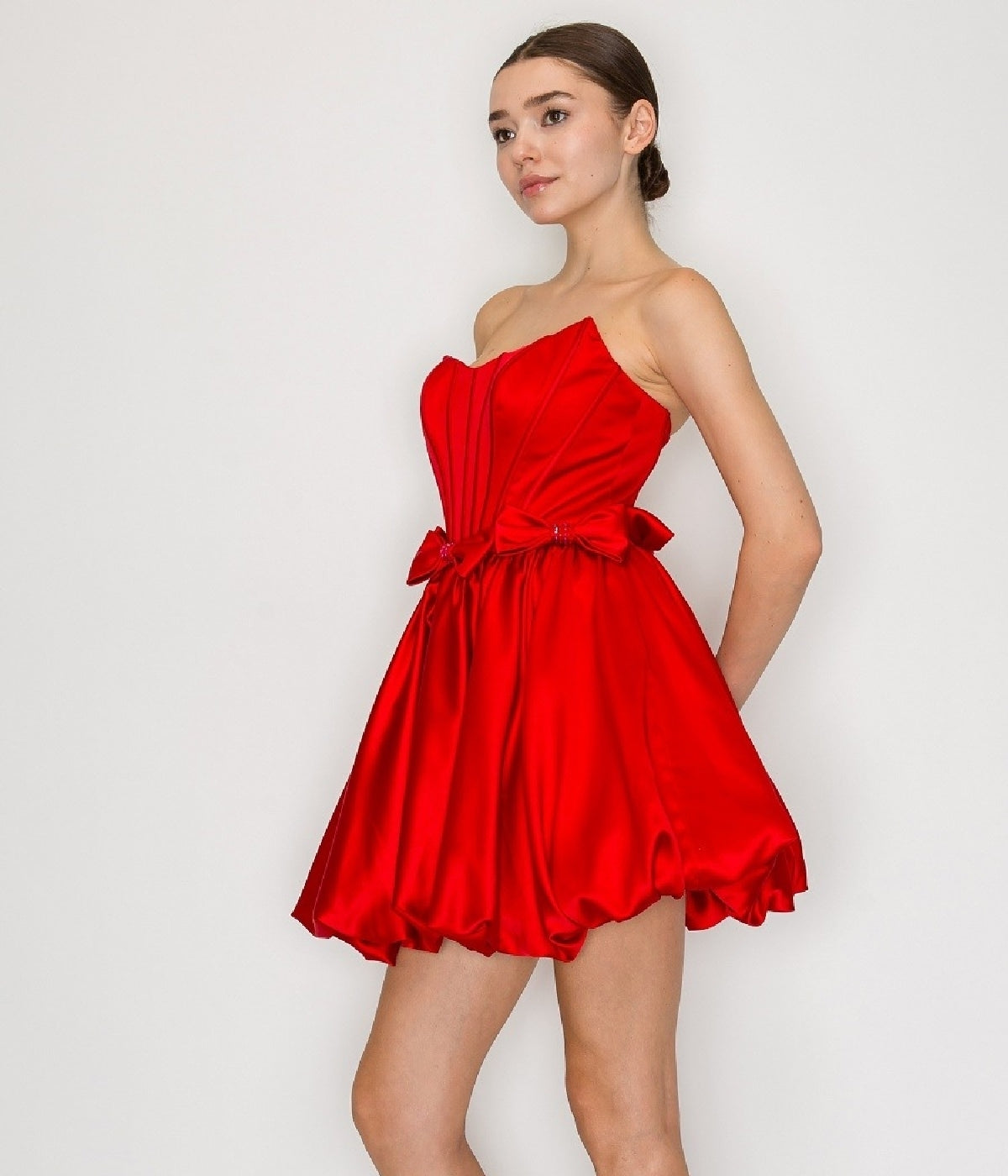 A woman wears the Strapless Short Bubble Party Dress: Poly USA 9950 in bright red satin, featuring a corset-style bodice and flared skirt, as she stands against a plain white background.